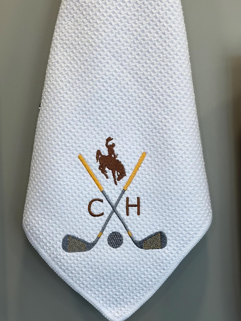 K&ouml;nnte beinhalten: Wei&szlig;es Golfhandtuch mit Waffelstruktur. Das Handtuch hat ein gesticktes Design mit gekreuzten Golfschl&auml;gern, einem Golfball und einer Cowboy-Silhouette auf einem Pferd. Die Initialen "C H" sind ebenfalls gestickt.