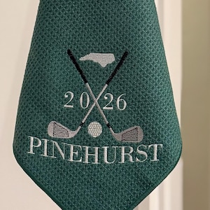 K&ouml;nnte beinhalten: Ein gr&uuml;nes Golfhandtuch mit Waffelstruktur. Das gestickte Design zeigt gekreuzte Golfschl&auml;ger, einen Golfball, die Zahl "20/26" und das Wort "PINEHURST" in Wei&szlig;.