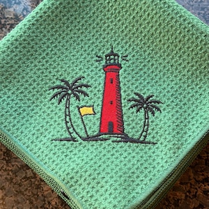 K&ouml;nnte beinhalten: Gr&uuml;nes Waffel-K&uuml;chentuch mit gesticktem Design. Das Design zeigt einen roten Leuchtturm mit schwarzer Umrandung, eine gelbe Flagge und zwei Palmen. Das Handtuch ist gefaltet und hat einen gr&uuml;nen Rand.