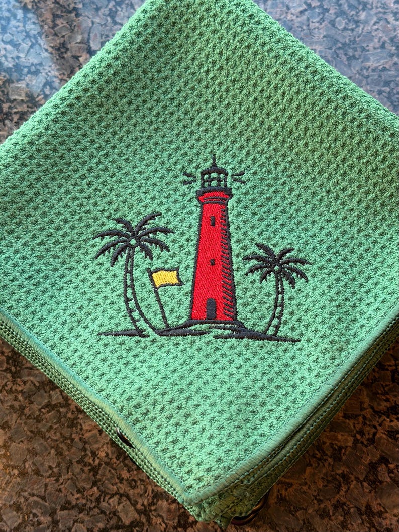 K&ouml;nnte beinhalten: Gr&uuml;nes Waffel-K&uuml;chentuch mit gesticktem Design. Das Design zeigt einen roten Leuchtturm mit schwarzer Umrandung, eine gelbe Flagge und zwei Palmen. Das Handtuch ist gefaltet und hat einen gr&uuml;nen Rand.