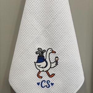 K&ouml;nnte beinhalten: Wei&szlig;es Golfhandtuch mit strukturierter Waffelwebung. Eine gestickte Cartoon-Gans tr&auml;gt Golfschl&auml;ger in einer blauen Tasche und ein Weinglas. Die Initialen "CS" sind blau mit Herzen gestickt.