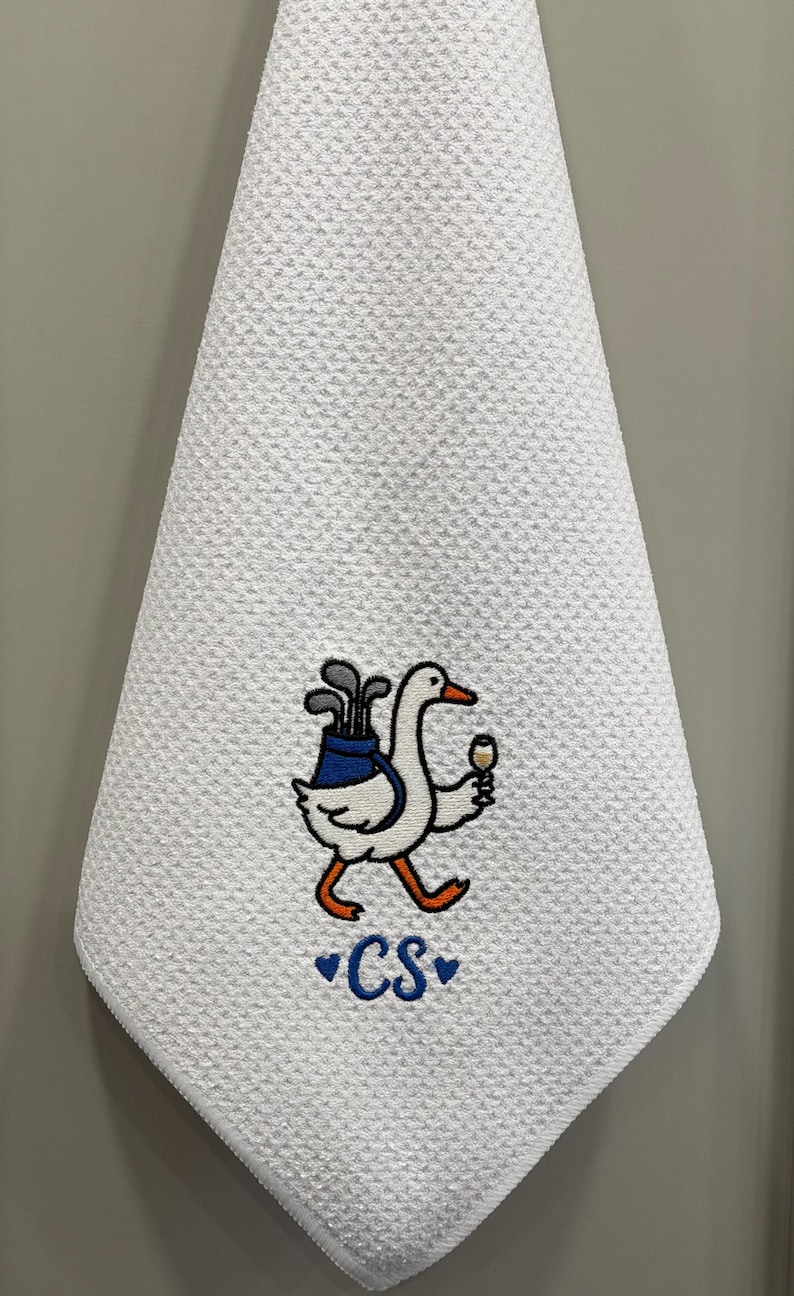 K&ouml;nnte beinhalten: Wei&szlig;es Golfhandtuch mit strukturierter Waffelwebung. Eine gestickte Cartoon-Gans tr&auml;gt Golfschl&auml;ger in einer blauen Tasche und ein Weinglas. Die Initialen "CS" sind blau mit Herzen gestickt.