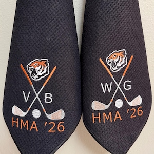 K&ouml;nnte beinhalten: Zwei schwarze Golfhandt&uuml;cher mit gestickten Designs. Jedes Handtuch hat ein Tigerkopf-Logo, gekreuzte Golfschl&auml;ger, einen Golfball und den Text "HMA '26". Ein Handtuch hat "VXB" und das andere "WXG".