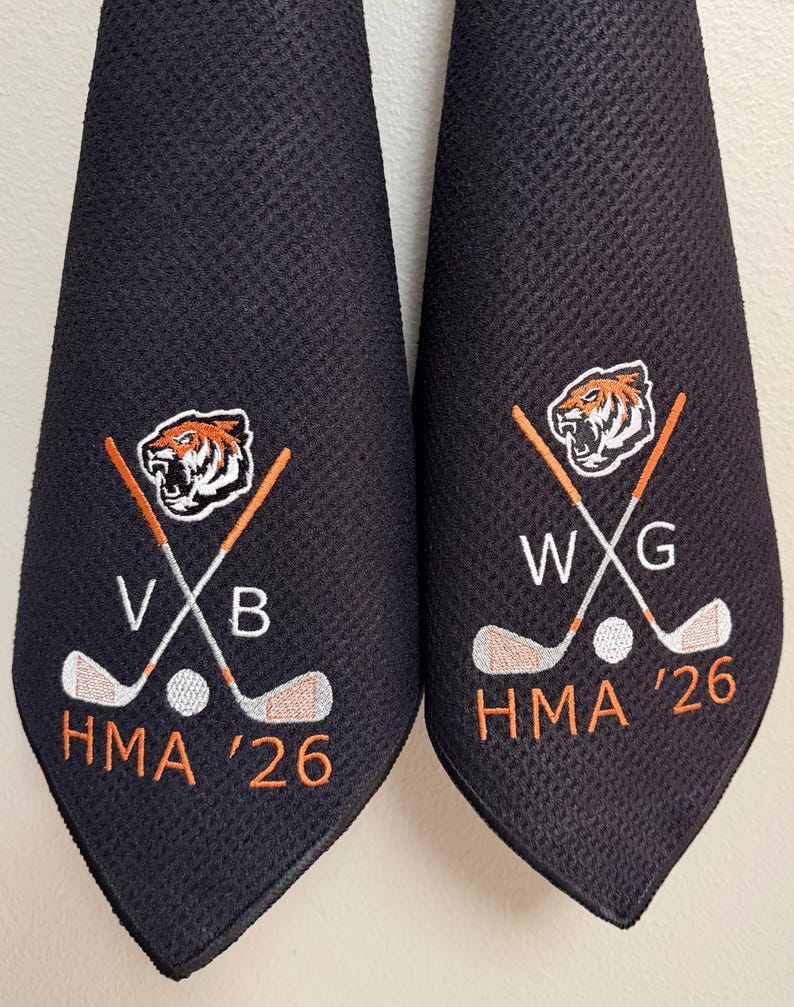 K&ouml;nnte beinhalten: Zwei schwarze Golfhandt&uuml;cher mit gestickten Designs. Jedes Handtuch hat ein Tigerkopf-Logo, gekreuzte Golfschl&auml;ger, einen Golfball und den Text "HMA '26". Ein Handtuch hat "VXB" und das andere "WXG".
