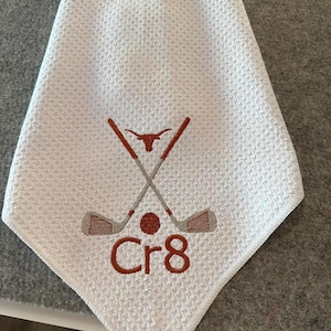 K&ouml;nnte beinhalten: Wei&szlig;es Golfhandtuch mit Waffelstruktur. Es hat ein gesticktes Design aus gekreuzten Golfschl&auml;gern, einem Golfball und dem Text "Cr8" in Braun. Ein Longhorn-Symbol befindet sich oben auf den gekreuzten Schl&auml;gern.