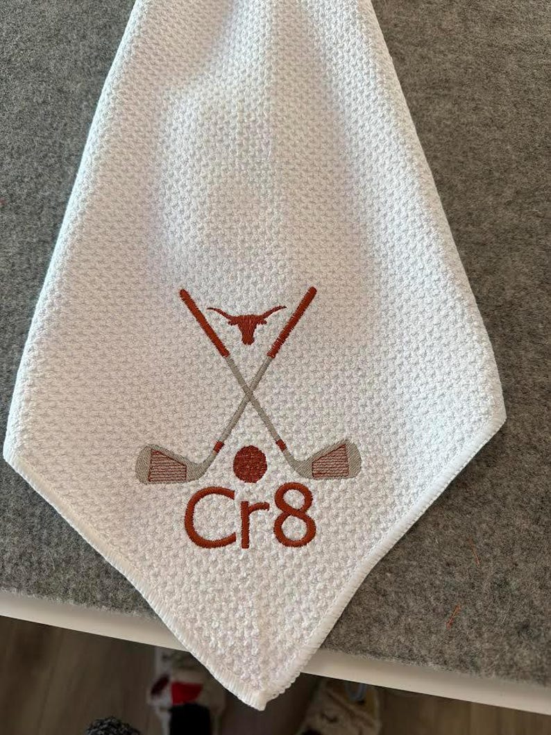 K&ouml;nnte beinhalten: Wei&szlig;es Golfhandtuch mit Waffelstruktur. Es hat ein gesticktes Design aus gekreuzten Golfschl&auml;gern, einem Golfball und dem Text "Cr8" in Braun. Ein Longhorn-Symbol befindet sich oben auf den gekreuzten Schl&auml;gern.