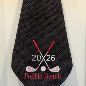 K&ouml;nnte beinhalten: Schwarzes Golfhandtuch mit einem gestickten Design. Das Design umfasst gekreuzte Golfschl&auml;ger, einen Golfball, die Zahlen "2026" und die Worte "Pebble Beach" in Rot.