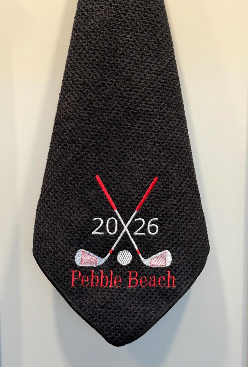 K&ouml;nnte beinhalten: Schwarzes Golfhandtuch mit einem gestickten Design. Das Design umfasst gekreuzte Golfschl&auml;ger, einen Golfball, die Zahlen "2026" und die Worte "Pebble Beach" in Rot.
