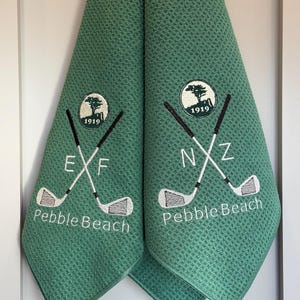 K&ouml;nnte beinhalten: Zwei gr&uuml;ne Golfhandt&uuml;cher mit Waffelstruktur. Jedes Handtuch hat ein gesticktes Logo mit gekreuzten Golfschl&auml;gern, dem Text "Pebble Beach" und dem Jahr 1919. Ein Handtuch hat die Buchstaben "EXF", das andere "NXZ".