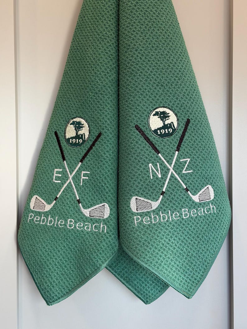 K&ouml;nnte beinhalten: Zwei gr&uuml;ne Golfhandt&uuml;cher mit Waffelstruktur. Jedes Handtuch hat ein gesticktes Logo mit gekreuzten Golfschl&auml;gern, dem Text "Pebble Beach" und dem Jahr 1919. Ein Handtuch hat die Buchstaben "EXF", das andere "NXZ".