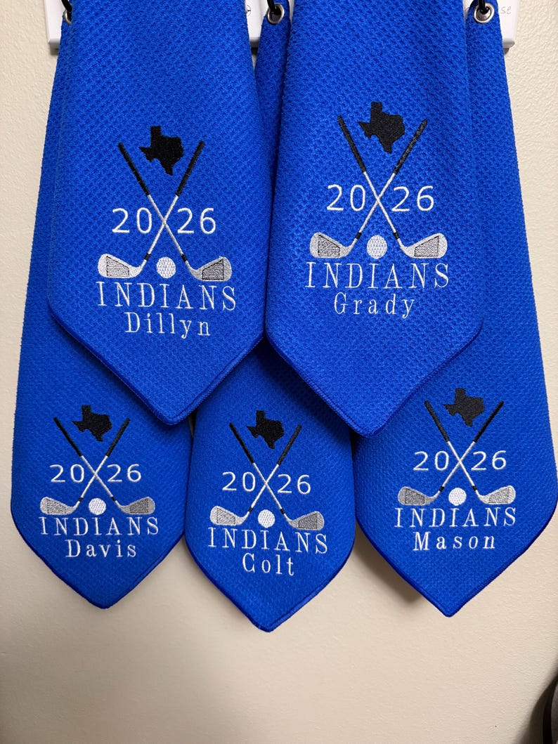 K&ouml;nnte beinhalten: Blaue Golfhandt&uuml;cher mit gestickten Designs. Jedes Handtuch zeigt den Text "20/26", gekreuzte Golfschl&auml;ger, einen Golfball und das Wort "INDIANS" zusammen mit einem Namen. Eine Texas-Umrisslinie ist ebenfalls gestickt.