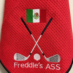 K&ouml;nnte beinhalten: Rotes Golfhandtuch mit schwarzem Rand, bestickt mit gekreuzten Golfschl&auml;gern, einem Golfball und dem Text "Freddie's ASS" in Wei&szlig;. Mit der mexikanischen Flagge. Ein lustiges Geschenk f&uuml;r einen Golfer.