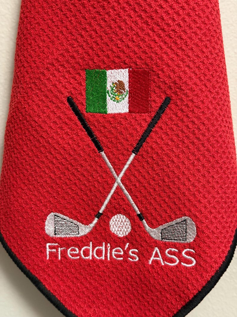 K&ouml;nnte beinhalten: Rotes Golfhandtuch mit schwarzem Rand, bestickt mit gekreuzten Golfschl&auml;gern, einem Golfball und dem Text "Freddie's ASS" in Wei&szlig;. Mit der mexikanischen Flagge. Ein lustiges Geschenk f&uuml;r einen Golfer.