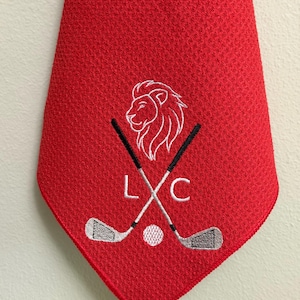K&ouml;nnte beinhalten: Eine rote Golfkrawatte mit strukturiertem Muster. Die Krawatte zeigt einen wei&szlig;en, gestickten L&ouml;wenkopf, gekreuzte Golfschl&auml;ger, einen Golfball und die Buchstaben "L" und "C".