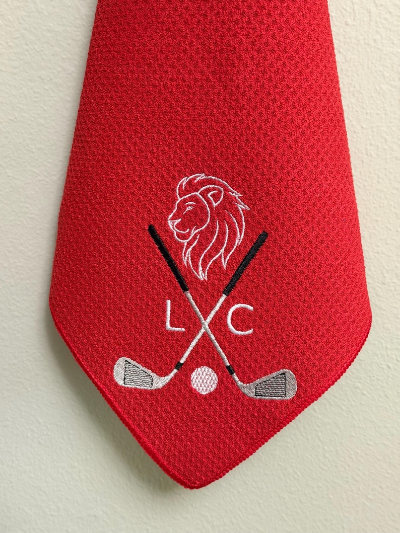 K&ouml;nnte beinhalten: Eine rote Golfkrawatte mit strukturiertem Muster. Die Krawatte zeigt einen wei&szlig;en, gestickten L&ouml;wenkopf, gekreuzte Golfschl&auml;ger, einen Golfball und die Buchstaben "L" und "C".
