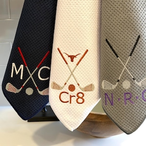 K&ouml;nnte beinhalten: Drei Golf-Handt&uuml;cher in Marine, Wei&szlig; und Grau. Jedes Handtuch zeigt gestickte Golfschl&auml;ger, einen Golfball und Initialen. Das marinefarbene Handtuch hat "MXC", das wei&szlig;e "Cr8" und das graue "NRC".