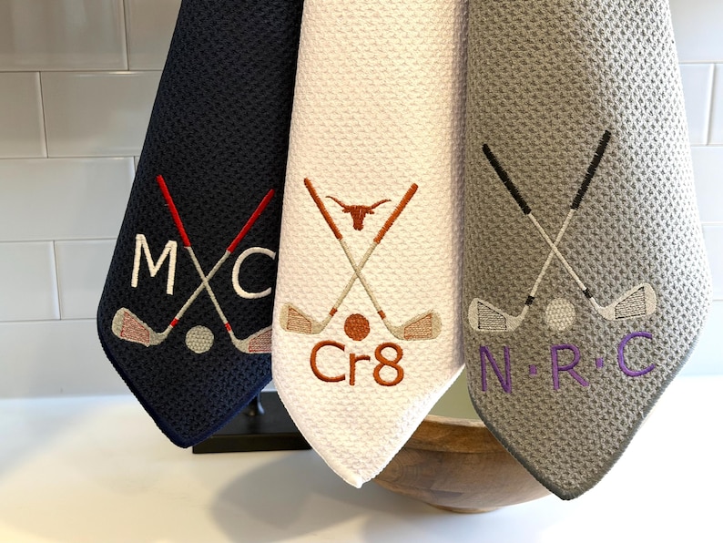 K&ouml;nnte beinhalten: Drei Golf-Handt&uuml;cher in Marine, Wei&szlig; und Grau. Jedes Handtuch zeigt gestickte Golfschl&auml;ger, einen Golfball und Initialen. Das marinefarbene Handtuch hat "MXC", das wei&szlig;e "Cr8" und das graue "NRC".