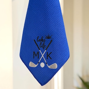 K&ouml;nnte beinhalten: Ein blaues Golfhandtuch mit den gestickten Worten "lake life" und den Initialen "MXK". Das Design umfasst gekreuzte Golfschl&auml;ger und einen Golfball. Das Handtuch h&auml;ngt.