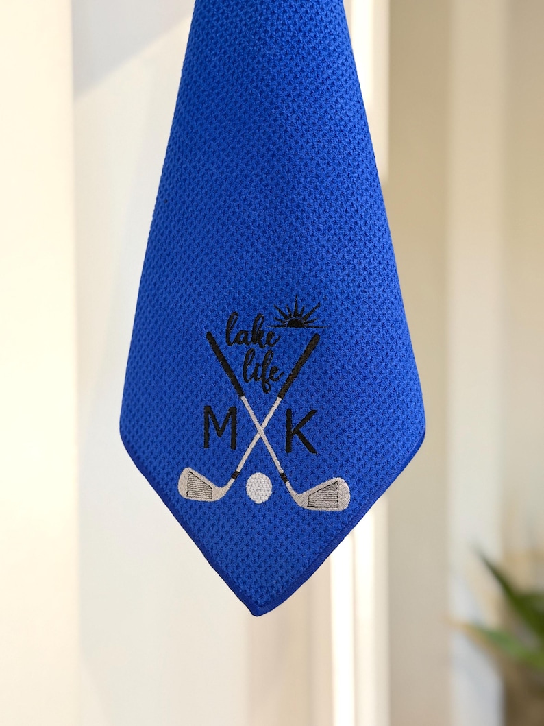 K&ouml;nnte beinhalten: Ein blaues Golfhandtuch mit den gestickten Worten "lake life" und den Initialen "MXK". Das Design umfasst gekreuzte Golfschl&auml;ger und einen Golfball. Das Handtuch h&auml;ngt.