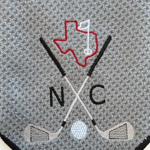 K&ouml;nnte beinhalten: Graues Golfhandtuch mit schwarzem Rand. Mit gesticktem Design aus gekreuzten Golfschl&auml;gern, einem Golfball und dem roten Umriss von Texas mit einer wei&szlig;en Flagge. Die Buchstaben "N" und "C" sind ebenfalls gestickt.