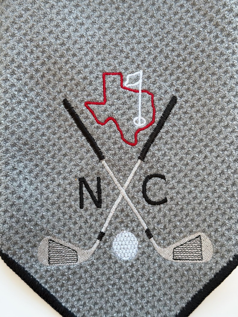 K&ouml;nnte beinhalten: Graues Golfhandtuch mit schwarzem Rand. Mit gesticktem Design aus gekreuzten Golfschl&auml;gern, einem Golfball und dem roten Umriss von Texas mit einer wei&szlig;en Flagge. Die Buchstaben "N" und "C" sind ebenfalls gestickt.