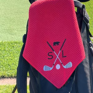 K&ouml;nnte beinhalten: Ein rotes Golftuch mit strukturierter Oberfl&auml;che, das an einer Golftasche h&auml;ngt. Das Tuch hat ein gesticktes Design mit gekreuzten Golfschl&auml;gern, einem B&auml;ren, einem Golfball und den Buchstaben "SXL" in Wei&szlig;.