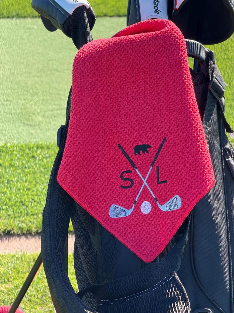 K&ouml;nnte beinhalten: Ein rotes Golftuch mit strukturierter Oberfl&auml;che, das an einer Golftasche h&auml;ngt. Das Tuch hat ein gesticktes Design mit gekreuzten Golfschl&auml;gern, einem B&auml;ren, einem Golfball und den Buchstaben "SXL" in Wei&szlig;.