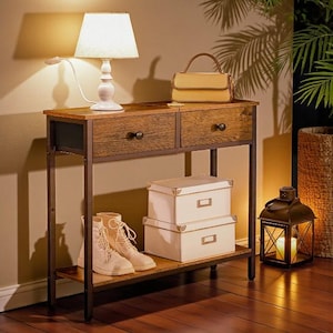May include: A wooden console table with two drawers, a lower shelf, and a metal frame. A white lamp, a tan handbag, two white storage boxes, and a pair of cream-coloured boots are on the table. A lit lantern is on the floor.