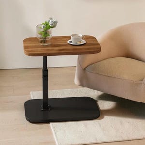 Puede incluir: Una mesa auxiliar de altura ajustable con una superficie rectangular de madera de esquinas redondeadas, sostenida por un poste de metal negro y una base plana negra. Un florero de vidrio transparente con follaje verde y una taza blanca.