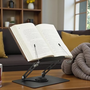 May include: A black metal book stand supports an open book on a wooden table. The stand features a rectangular base and adjustable arms. A grey blanket and yellow cushion are in the background.