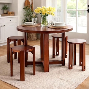 May include: A round, dark wood dining table with a central pedestal base and four matching round stools. The table is set with stacks of white plates, clear glasses, and a glass vase filled with bright yellow daffodils on a woven placemat.