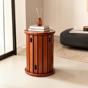 May include: A cylindrical wooden side table with vertical slats and oval cutouts. The table has a flat circular top surface holding two stacked white books and a small amber glass bottle containing thin wooden reed diffuser sticks.
