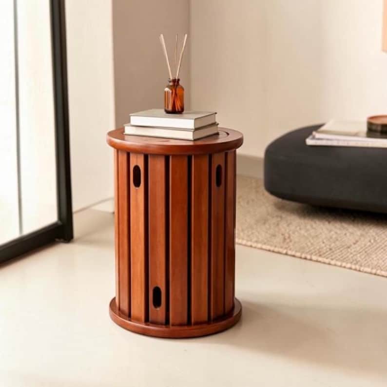 May include: A cylindrical wooden side table with vertical slats and oval cutouts. The table has a flat circular top surface holding two stacked white books and a small amber glass bottle containing thin wooden reed diffuser sticks.