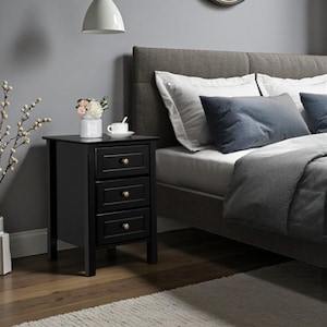 Pode incluir: Uma mesa de cabeceira preta com três gavetas e puxadores dourados. A mesa de cabeceira está ao lado de uma cama com cabeceira cinza e roupa de cama branca. Um pequeno vaso de flores e uma chávena e pires estão em cima.