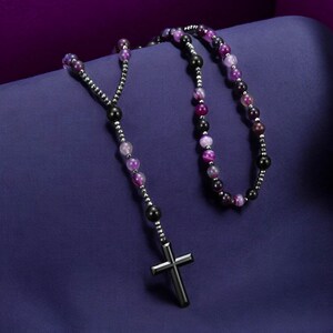 May include: A rosary necklace featuring a silver cross pendant and beads in shades of purple, black, and silver. The beads are strung on a silver chain, creating a religious accessory.