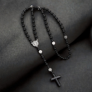Puede incluir: Un rosario negro con un colgante de cruz. El collar presenta peque&ntilde;as cuentas negras redondas y medallones religiosos plateados. La cruz es de forma rectangular simple.