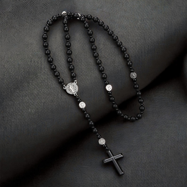Puede incluir: Un rosario negro con un colgante de cruz. El collar presenta peque&ntilde;as cuentas negras redondas y medallones religiosos plateados. La cruz es de forma rectangular simple.