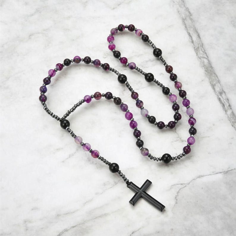 May include: A rosary featuring purple and black beads, connected by a dark metal chain. A black cross is suspended at the base. The beads display a marbled effect, showcasing hues of purple and black. The rosary is a religious item.