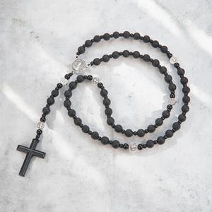 Puede incluir: Rosario negro con cuentas de piedra volc&aacute;nica y detalles plateados. El rosario presenta un crucifijo y elementos decorativos met&aacute;licos. Las cuentas est&aacute;n espaciadas uniformemente, y el dise&ntilde;o es sim&eacute;trico. El rosario se muestra sobre una superficie clara.