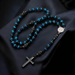 Puede incluir: Un collar de rosario con cuentas de ojo de tigre azul y detalles de piedra de lava negra. El collar incluye un medallón plateado y un colgante de cruz negra. Las cuentas están ensartadas en un cordón oscuro.