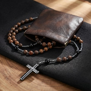 Puede incluir: Un rosario con cuentas de madera marrón, pequeñas cuentas de color gris metálico y cuentas negras más grandes, que termina en un colgante de cruz de metal oscuro y liso. El collar está parcialmente guardado en una pequeña bolsa de cuero marrón.