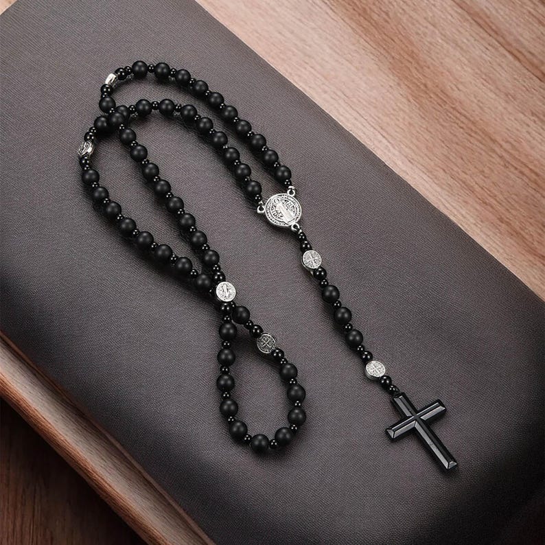 Puede incluir: Rosario de cuentas negras con una cruz plateada y medallones religiosos circulares. El rosario se muestra sobre una superficie gris oscuro con un fondo de madera. La cruz tiene un dise&ntilde;o simple y moderno.