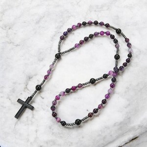May include: A rosary featuring a black cross pendant and beads in shades of purple, black, and grey. The beads are strung on a dark grey chain, a religious item.