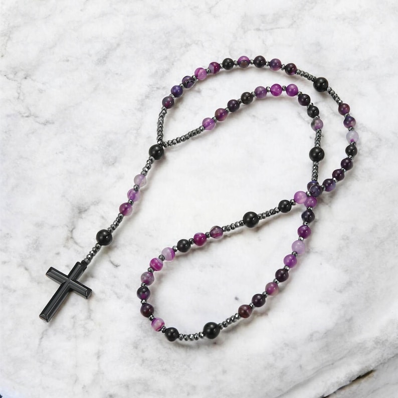 May include: A rosary featuring a black cross pendant and beads in shades of purple, black, and grey. The beads are strung on a dark grey chain, a religious item.