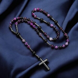 May include: A rosary necklace featuring a silver cross pendant and beads in shades of purple, black, and white. The beads are strung on a silver chain, displayed on dark blue fabric.