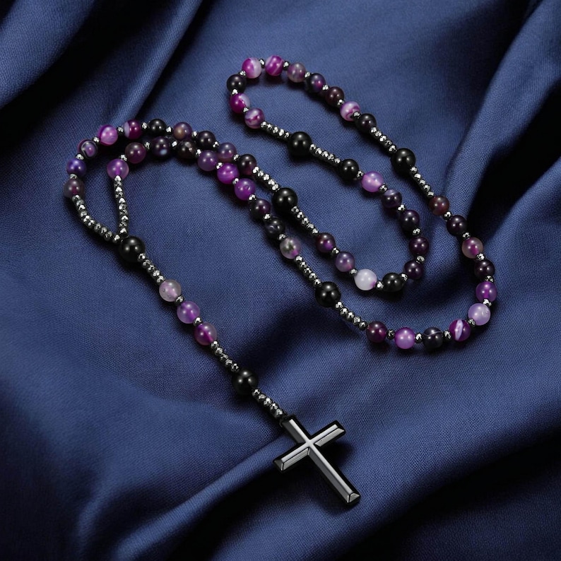 May include: A rosary necklace featuring a silver cross pendant and beads in shades of purple, black, and white. The beads are strung on a silver chain, displayed on dark blue fabric.