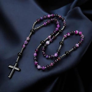 May include: A rosary featuring purple and black beads and a silver cross. The beads are a mix of colours, including shades of purple and black. The cross is a simple, modern design.