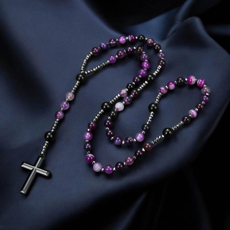 May include: A rosary featuring purple and black beads and a silver cross. The beads are a mix of colours, including shades of purple and black. The cross is a simple, modern design.