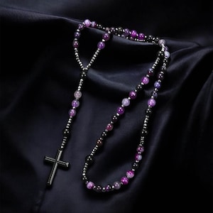 Puede incluir: Un collar de rosario con un colgante de cruz de metal oscuro y una cadena de cuentas moradas y negras. Las cuentas están intercaladas con pequeñas cuentas de color plateado. El collar se muestra sobre un fondo oscuro y texturizado.