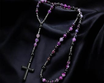 May include: A rosary necklace featuring a dark metal cross pendant and a strand of purple and black beads. The beads are interspersed with smaller silver-colored beads. The necklace is displayed on a dark, textured background.